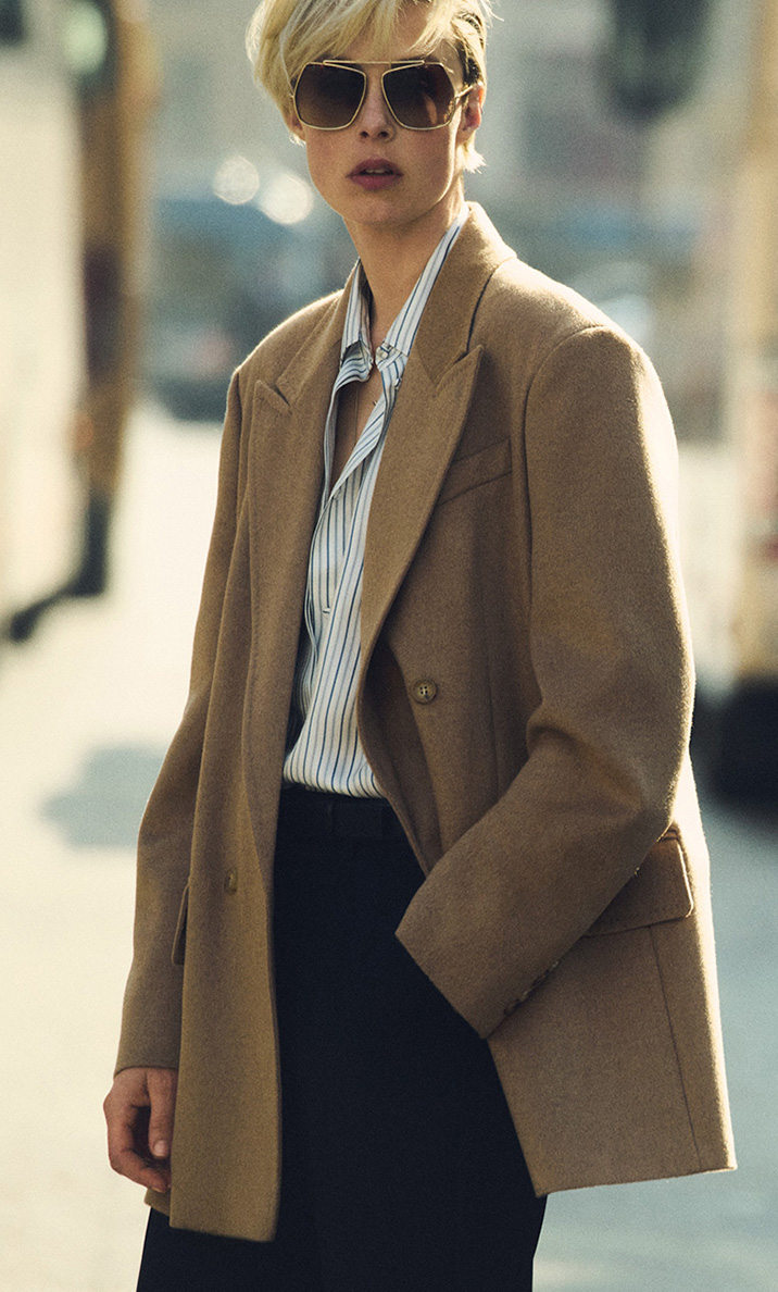 Das Model trägt den doppelreihigen Blazer Olimpia von Max Mara im Farbton Kamel zu dunkler Brille und Bluse im urbanen Kontext.
